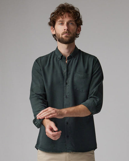 A person wearing a dark olive button-down shirt with buttoned sleeves, standing against a grey background.